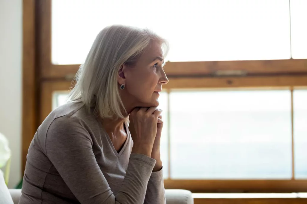 Donna matura seduta su un divano, guarda fuori dalla finestra con sguardo triste e sconvolto, che rappresentano il concetto di pensione di reversibilità
