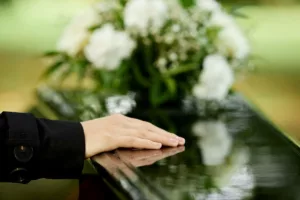 Mano su una bara lucida con fiori bianchi, rimborso delle spese funerarie fino a 294 euro.